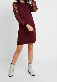 Burgundy klänning med transparenta, veckade långa ärmar som pryds av blommig broderi. Modellen matchar den med texturerade vita ankelstövlar i ormskinnsmönster.