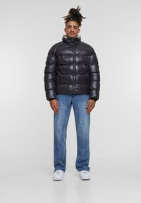 Zwarte pufferjack met glanzende afwerking, hoge kraag en rits aan de voorkant. Gecombineerd met blauwe rechttoe rechtaan jeans en witte sneakers.