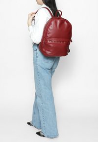 Un sac à dos en simili cuir rouge avec une forme arrondie, une poche avant zippée et des bretelles réglables, porté de manière décontractée avec un jean bleu clair.