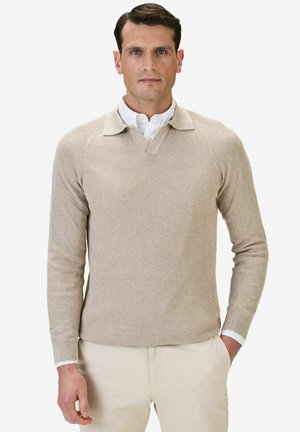 Beige ribbad stickad tröja med V-halsringning och krage, som bärs över en vit skjorta med krage. Raglanärmar och rak fåll.