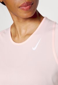 Vaaleanpunainen urheilutanktoppi, joka on valmistettu sileästä kankaasta ja jossa on pyöreä kaula-aukko sekä valkoinen Nike-logo vasemmassa rinnassa.