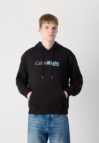Black cotton hoodie with front pocket, drawstring hood, and white and light blue "Calvin Klein NEW YORK" logo on the chest. Casual fit.