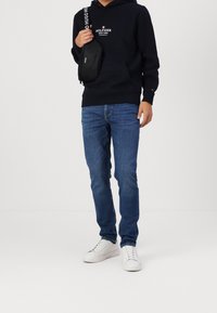 Sudadera con capucha negra y logo blanco en el pecho, jeans azules y zapatillas blancas. Pequeño bolso cruzado negro con marca blanca.