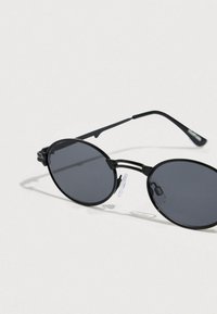 Στρογγυλά γυαλιά ηλίου με μαύρα πλαίσια, σκουρόχρωμους φακούς και διαφανείς επιθέματα στη μύτη. Διαθέτουν κομψές μεταλλικές λεπτομέρειες και ελαφρύ σχεδιασμό.