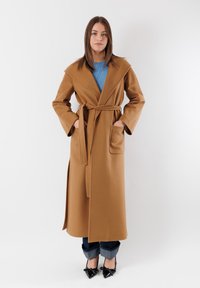 Cappotto lungo, marrone, con cintura, ampi risvolti, tasche a toppa e taglio dritto. Indossato su un top blu e abbinato a jeans scuri e tacchi neri.