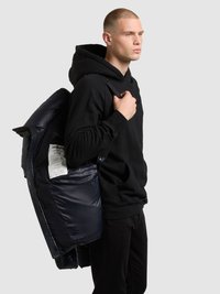 Črna kapucar s puloverjem, ki ga nosi oseba, drži mornarsko modro puhovko z bleščečim zaključkom in vidno etiketo znotraj.