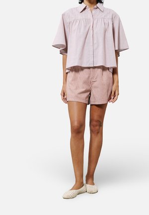 Donna con una camicetta rosa chiaro a maniche corte con bottoni, pantaloncini a righe rosse e bianche e scarpe beige intrecciate senza lacci, in piedi davanti a uno sfondo bianco.