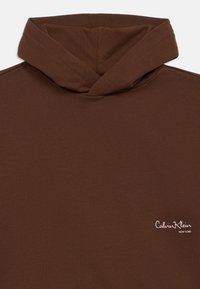 Καφέ φούτερ με σχέδιο πτυχής στον λαιμό, κατασκευασμένο από μαλακό ύφασμα. Διαθέτει λευκό λογότυπο "Calvin Klein New York" στο κάτω δεξί μέρος.