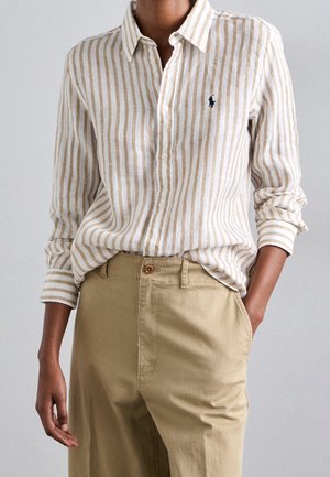 Person iført beige og hvidt vertikalt striped skjorte stukket ind i khakibukser med den ene hånd i lommen mod en ensfarvet baggrund.