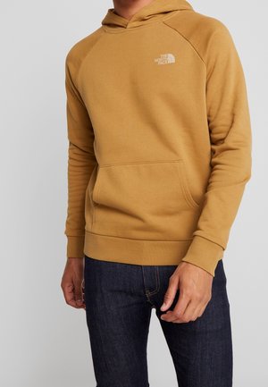 Mann trägt einen senfgelben The North Face Hoodie mit Fronttasche und dunkelblauen Jeans vor einem hellgrauen Hintergrund.