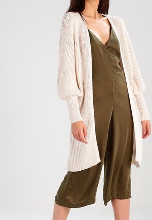 Ljust rosa stickad cardigan med långa puffärmar, bärs över en olivgrön jumpsuit. Tyget har en strukturerad, öppen väv.