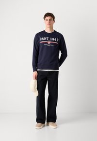 Marineblauwe sweatshirt met wit en rood "GANT 1949" logo, gedragen met zwarte wijde broek en beige sneakers, terwijl hij een lichtbeige pet vasthoudt.