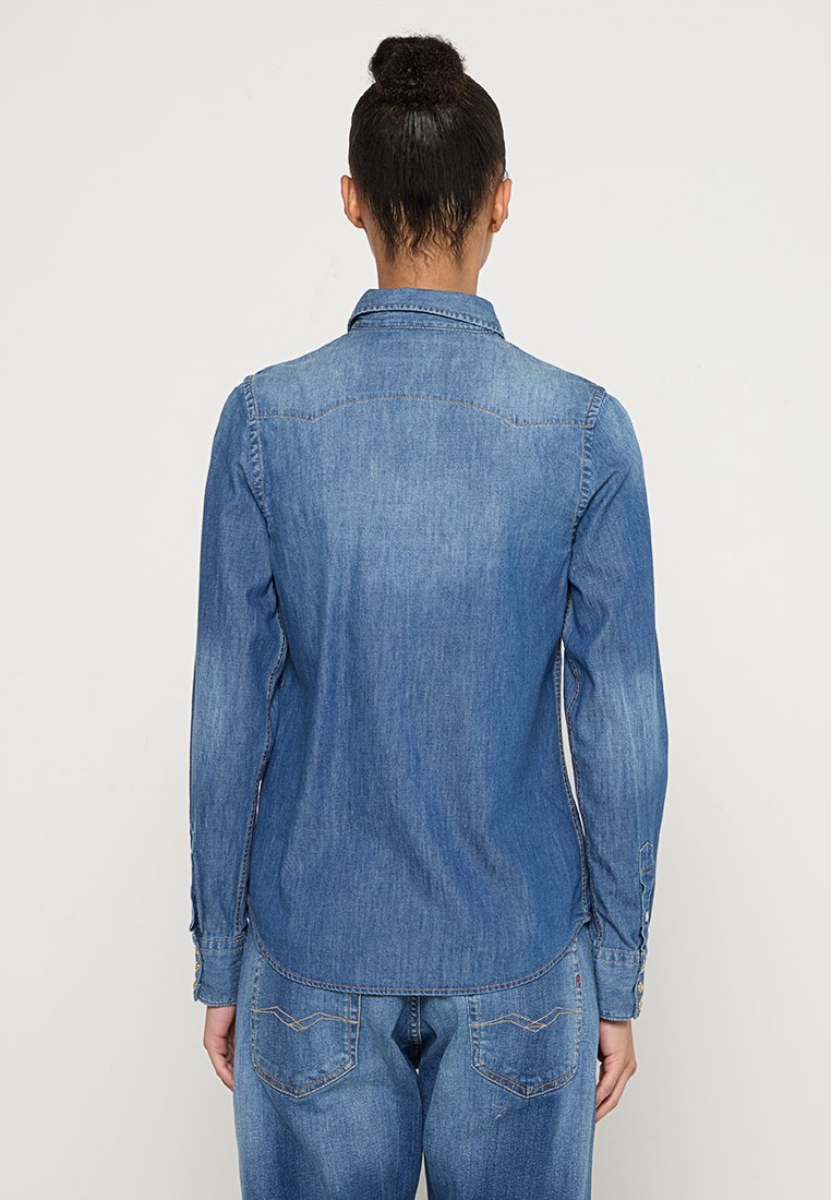 Camisa de denim en azul medio, con diseño de botones y cuello, mangas largas, detalle en la yugo trasera y puños con botones. Textura suave, corte casual.