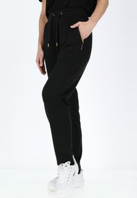 Zwarte joggingbroek van zachte stof, met een trekkoord in de taille, ritszakken aan de zijkant en een taps toelopende pasvorm. G gedragen met witte sneakers.