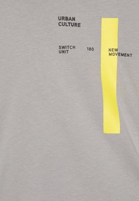 Una camiseta gris claro presenta texto negro y una franja amarilla vertical. El material parece suave con un diseño de ajuste clásico.