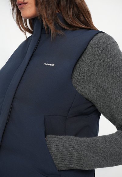 Námornícky puffer vest s vysokým golierom a logom na hrudi. Šedé textúrované sveterové rukávy sa rozširujú od otvorov na rukávy. Obsahuje bočné vrecká.