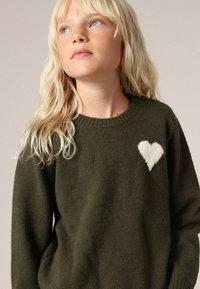 Donkergroene gebreide trui met een ronde hals en een witte hartaccent aan de linkerkant. Zachte textuur en lange mouwen.