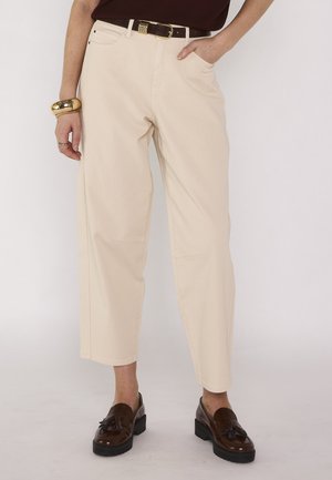 Femme portant un pantalon crème large et court, ceinture foncée, haut sombre, mocassins marron à pompons, bracelet en or et bague, debout avec une main dans la poche.