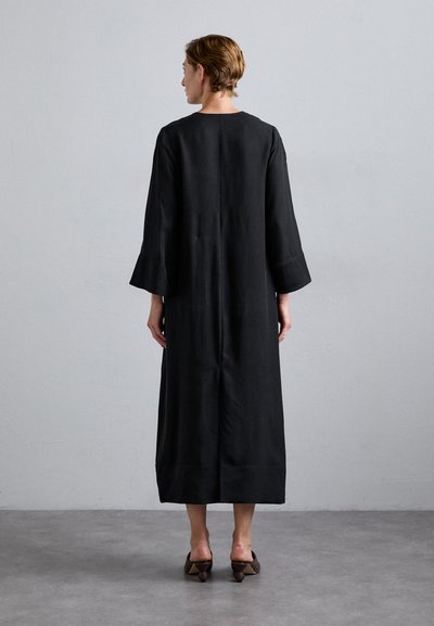 Black long dress with wide sleeves, simple design, and a straight cut, paired with brown high-heeled shoes. Background is plain.