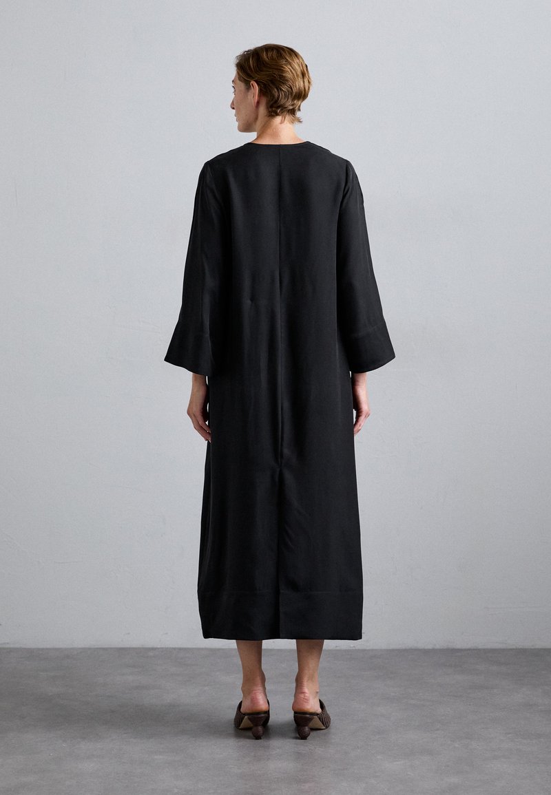 Schwarzes langes Kleid mit weiten Ärmeln, schlichtem Design und geradem Schnitt, kombiniert mit braunen High Heels. Der Hintergrund ist einfarbig.