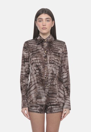 Donna con capelli lunghi e scuri, indossa una camicia marrone e beige con motivo a bottoni e maniche lunghe abbinata a pantaloncini, su uno sfondo semplice.