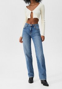 Suéter corto de canalé crema con escote en V, combinado con jeans de pierna recta azul de cintura alta y botas negras de tobillo.
