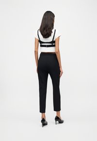 Γυναίκα με μακριά σκούρα μαλλιά στέκεται γυρισμένη πλάτη, φοράει ένα λευκό κοντό τοπ με μαύρες τιράντες, μαύρο παντελόνι tailored και μαύρα ψηλοτάκουνα παπούτσια.