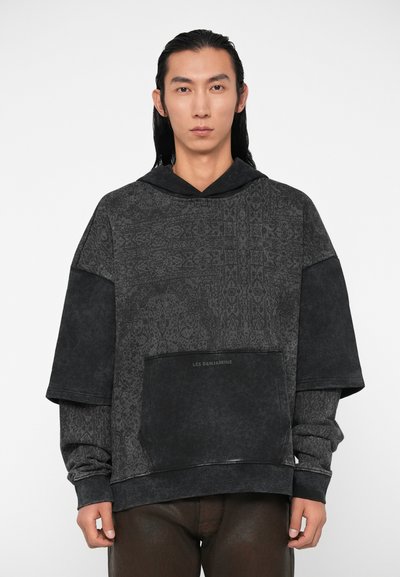 Felpa nera oversize con texture a motivi, dotata di una grande tasca frontale e cappuccio. Il materiale sembra morbido con una lucentezza attenuata.