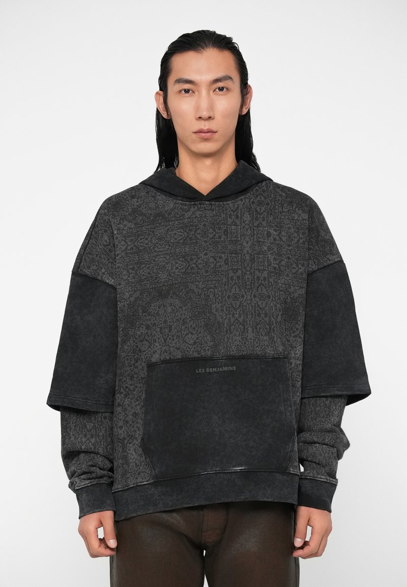 Prevelik crni dukserica s uzorkovanom teksturom, koja ima veliki prednji džep i kapuljaču. Materijal izgleda mekan s suptilnim sjajem.