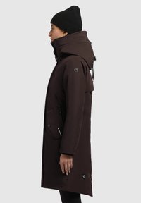 Chaqueta parka marrón con un collar alto, bolsillos con cremallera y capucha ajustable. Presenta un logo triangular en el lado inferior. Tela texturizada.