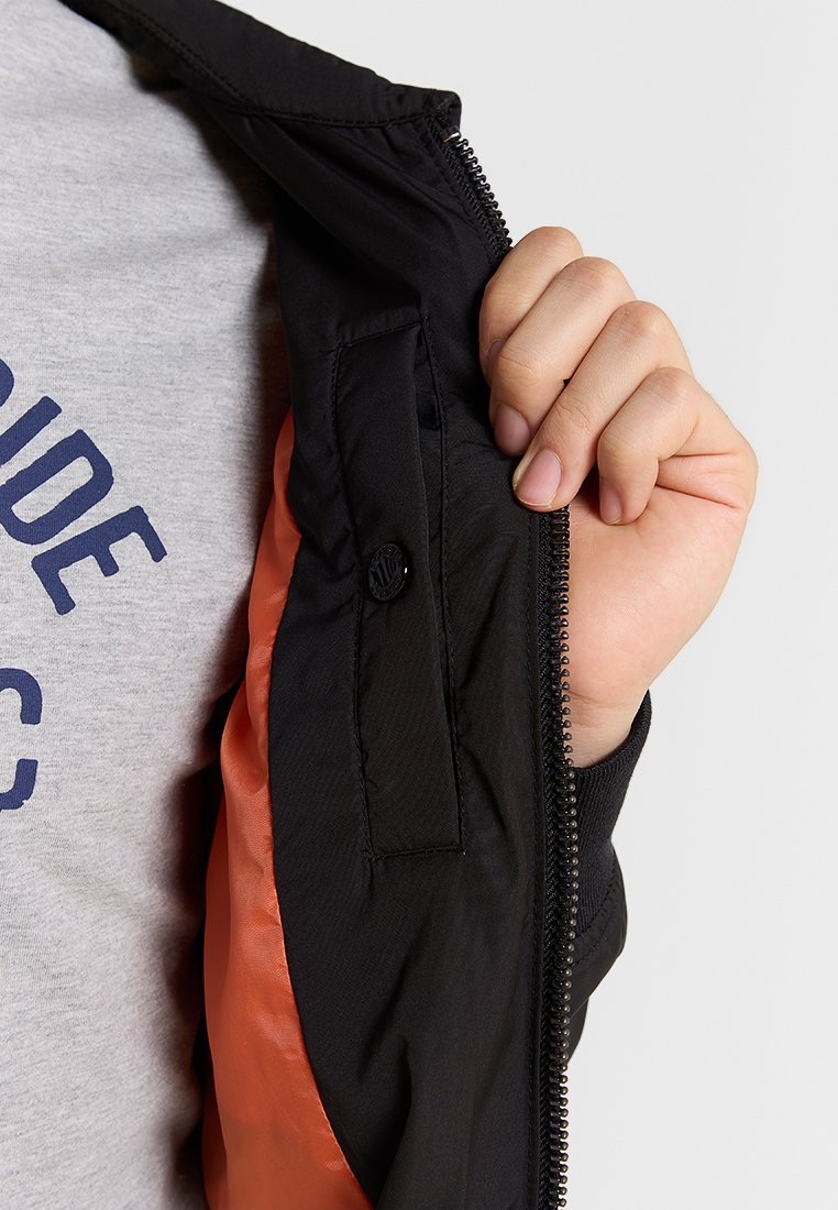 Veste noire avec une doublure orange, fermeture éclair, et une poche à bouton à l'intérieur. Texture lisse et matériau léger visibles.