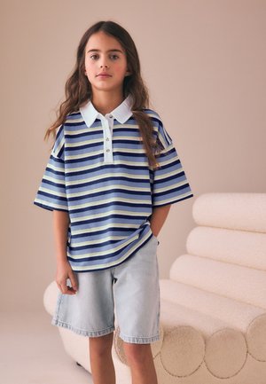 Ung pige med langt hår iført en blåstribet polo og lyse denimshorts står ved siden af en beige tekstureret sofa.