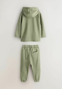 Oliwkowa bluza z kapturem, z przeszyciami na rękawach. Dopasowane spodnie dresowe z elastycznym pasem i logo na tylnej kieszeni.