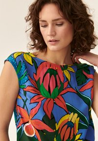 Floral gemusterte Bluse in lebhaften Farben, mit großen roten und gelben Blumen auf einem blauen Hintergrund, mit glatter Textur und Schulterdetails.