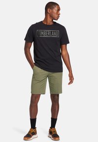 Sort bomulds t-shirt med "TIMBERLAND" tekst på brystet, olivengrønne shorts og brune sportsko med sorte detaljer og gule snørebånd.