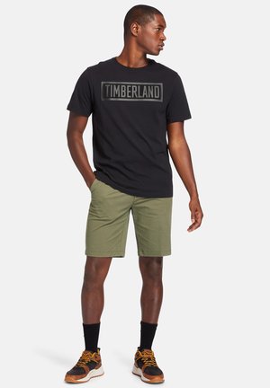 T-shirt en coton noir avec le texte "TIMBERLAND" sur la poitrine, shorts vert olive et chaussures de sport marron avec accents noirs et lacets jaunes.