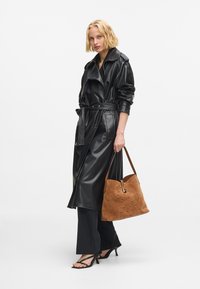 Černý kabát z umělé kůže s páskem v pase, spárovaný s měkkou hnědou semišovou kabelkou s jemným vzorem, úzkými černými kalhotami a sandály na vysokém podpatku.