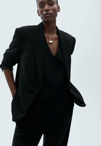 Črni eleganten blazer preko črnega telovnika, ki ima ovratnik in tri gumbe. Model nosi zlat medaljon, na enostavni ozadju.