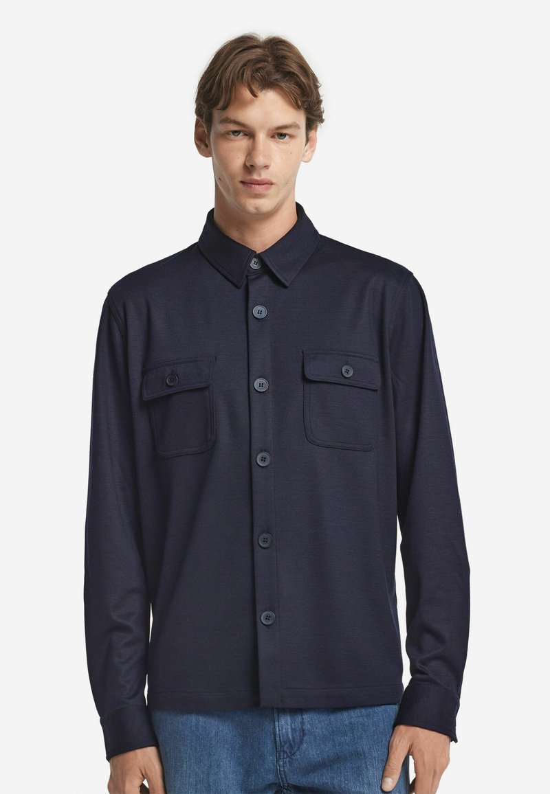 Granatowa koszula z długim rękawem, z kołnierzykiem, dwoma przednimi kieszeniami i zapięciem na guziki. Wykonana z gładkiego materiału o klasycznym kroju.