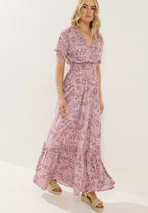 Kvinde iført en lyserød blomsterprint maxi-kjole med korte puffærmer og beige platformsandaler, stående foran en ensfarvet lys baggrund.