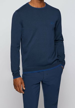 Homme portant un pull bleu marine à manches longues avec un logo discret sur la poitrine, assorti d’un pantalon bleu marine, main dans la poche.