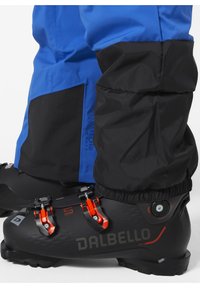 Černé lyžařské boty s oranžovými detaily, s texturovaným povrchem, vedle modrých sněhových kalhot s černým nápletem a logem.