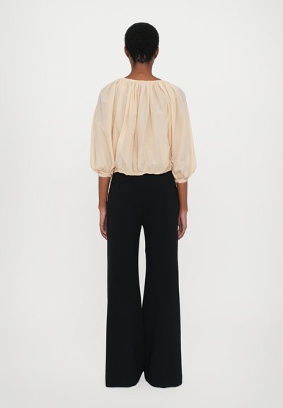 Pfirsichfarbene Bluse mit Ballonärmeln und elastischen Bündchen, kombiniert mit weiten schwarzen Hosen. Glatter Stoff, leichtes Design, minimales Detail.