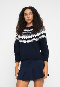 Junge Frau mit lockigem Haar, die einen navyfarbenen Pullover mit weißen geometrischen Mustern und einen navyfarbenen Faltenrock trägt, steht vor einem weißen Hintergrund.