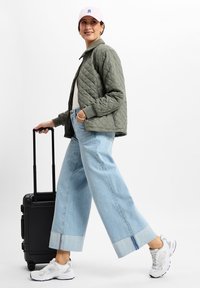 Gequilted olijfgroene jas, lichtgrijze shirt, brede lichtblauwe jeans met opgerolde zomen, witte sneakers, en een roze pet met blauw logo. Zwarte koffer.