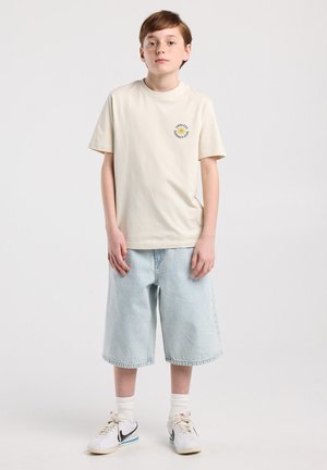Garçon portant un t-shirt beige "Endless Summer Club", un short en jean bleu clair, des chaussettes blanches et des baskets blanches Nike, debout devant un fond uni.