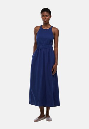 Femme portant une robe midi bleu marine sans manches avec une taille cintrée et un tissu texturé, debout devant un fond clair uni.