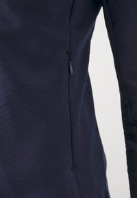 Haut ajusté bleu marine en tissu doux, avec une fermeture éclair sur le côté et une texture lisse avec un léger éclat.