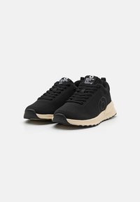 Des baskets noires avec une tige en maille texturée, un intermédiaire de couleur crème et une semelle extérieure en caoutchouc noir ; présente un logo sur la languette et des détails sur le côté.