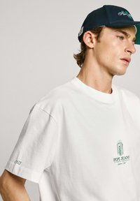 Camiseta de algodón blanca con mangas cortas, que presenta un logo bordado verde en el pecho y "1973" cosido en la manga.